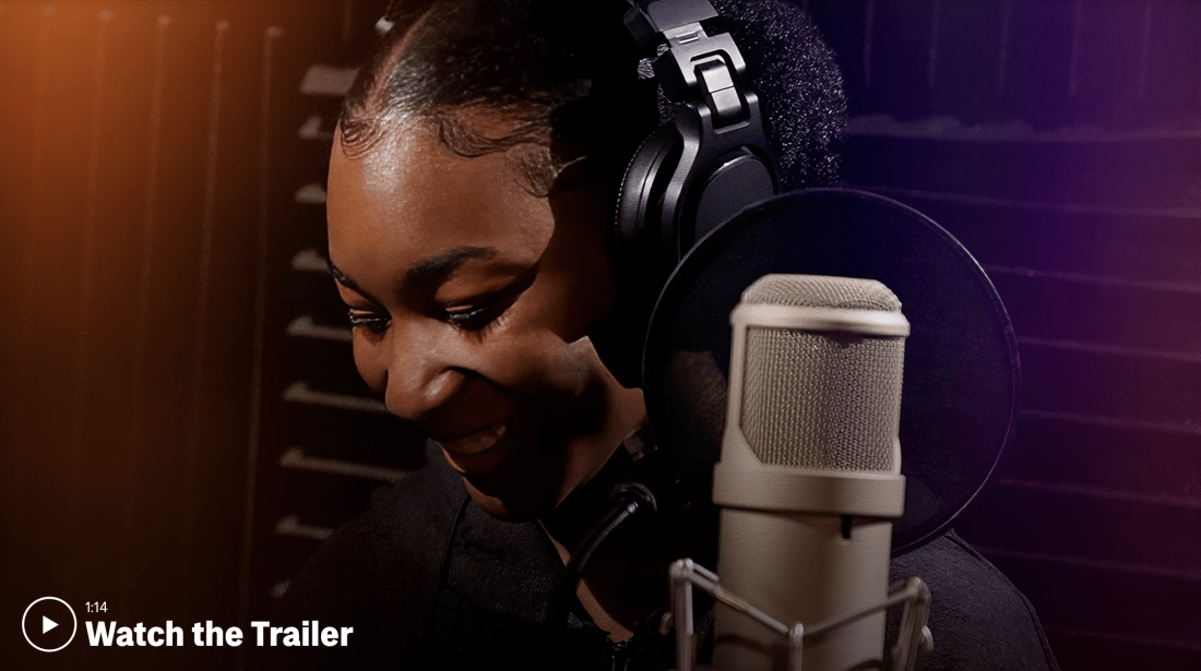 Photo of a student smiling while wearing headphones in front of a studio recording microphone with a white play button and the text "Watch the Trailer"